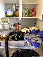 Wide view showing nested plastic bowls, plastic glasses, white disposable plates, bbq grill pan, cooking utensils, and bags on top of a washing machine close to white cabinetry.