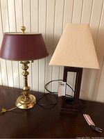 Both table lamps side by side on a wooden surface against a paneled wall. Brass lamp with maroon metal shade on the left and wood Bombay style lamp with paper shade on the right.