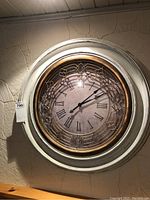 Front view of ornate shabby chic wall clock showing white distressed frame, gold decorative metal overlay, and Roman numeral clock face.