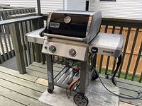 Full view of Weber Genesis II 2-burner gas barbecue showing black lid with thermometer, control knobs, side tables, and open stand with wheels.