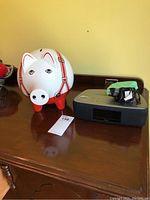 Photo showing the white ceramic piggy bank with red strap details and an iHome clock radio docking station with green tape on top, both placed on a wooden surface.