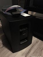 Front view of the Bose subwoofer with a remote control placed on top inside a plastic bag