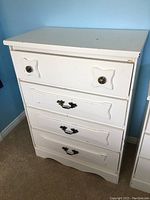 Front view of the white wood chest of drawers showing the four drawers and ornate metal knobs and handles with some wear.