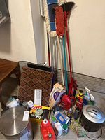 Photo showing assembled lot of cleaning equipment and supplies including mops, broom, Swiffer, rubbish bin, shredder, spray cleaners, and garbage bags.