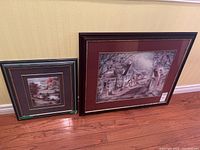 Photo showing two framed and matted prints leaning against a wall on a wood floor. The larger print features a village road scene and the smaller print shows a cottage on water.