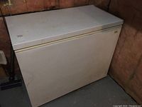 Full view of a white chest freezer with slight dirt and scratches, placed against basement wall with concrete floor.