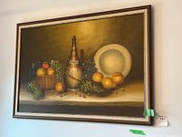 Original oil painting on canvas showing a detailed still life with fruit basket, wine bottle wrapped in straw, white plate, grapes, apples, pears, and cherries. Framed and hanging on a wall.