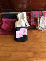 Photo of several folded tablecloths and napkins laid on the floor including burgundy linen, white lace, red green plaid Christmas, and tan lace patterned napkins.