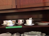 Shelf holding six tea cups each paired with saucers and two decorative plates on the right side. Includes various patterns and styles.