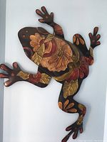 Full view of metal frog wall art mounted on wall, showing floral pattern in gold, copper, and red hues.