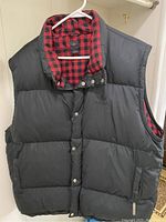 Front view of black Adidas puffy vest hanging on a white hanger showing puffy quilted design with snap front buttons.