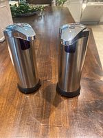 Two stainless steel battery operated automatic soap dispensers standing upright on a wooden table, shown from front angle.