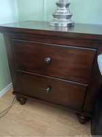 Front view of a dark brown wooden bedside table showing two closed drawers, round metal knobs and bun feet