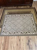 Two brown rugs displayed on wooden floor, smaller rug with diamond and floral border pattern placed on top of larger rug with speckled and decorative border pattern.