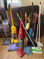 Photo of various cleaning tools including upright vacuum cleaners, mops, and dusters arranged on the floor against a dark brown sofa.