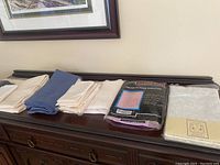 Seven fabric tablecloths in shades of cream and blue along with napkins laid out on a wooden surface. One blue round tablecloth visible folded among cream/beige linens.