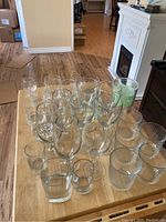 Top-down and side view showing the full grouping of assorted glassware on a wooden table including wine glasses, tumblers, shot glasses, and decorative patterned glasses.