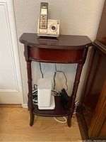 Full front view of wooden half-moon hallway table with drawer, top surface holding cordless phone and camera. Lower shelf holding networking devices.