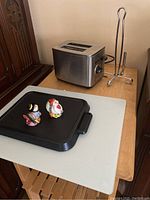 Photo showing a stainless steel two-slice toaster, metal paper towel holder, large glass cutting board on a wooden table, along with a black griddle pan and two turkey shaped salt and pepper shakers on top of the griddle.