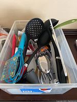 Various kitchen utensils including whisks, spoons, ladle, spatulas mainly in black, white, and blue colors inside a clear plastic bin.