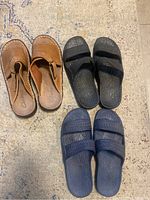 Three pairs of men's size 9 sandals laid flat on a rug floor: brown leather Clarks slip-ons, black dual strap slip-ons, navy blue dual strap slip-ons.