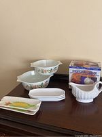 Photo of two Pyrex serving bowls with orange floral patterns, three rectangular corn-themed plates, a white ceramic gravy boat, and a boxed syrup and gravy server.