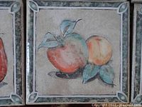 Close-up of a vintage ceramic tile showing a painted apple and peach with three leaves inside a decorative bordered square.