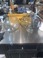 Both glass items shown on metallic surface: cake stand with dome cover on right, glass bubble bowl on left.