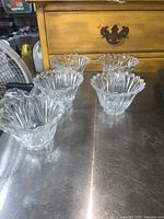 Five clear glass dessert dishes with scalloped edges arranged in two rows on a metal surface, showing their flower-like shape and thickness.