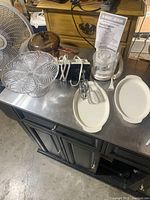 Photo showing the overall lot items including the cake stand, sauce pot with lid, handheld mixer, choppe, and plastic dishes displayed on metal surface