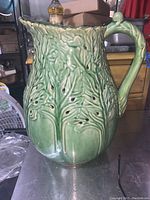 Side view of the green ceramic pitcher with raised leaf pattern, perforations, and handle shaped like a stem.