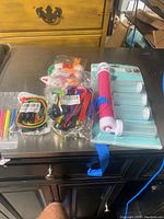 Photo showing assorted items on a tabletop including small bags with lanyards, colorful markers resembling syringes, a pink and white manual cake airbrush tool packaged with plastic tubes.