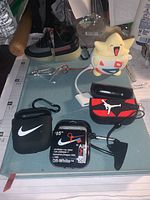 Five AirPod cases themed with Nike, Off-White, Air Jordan, small sneaker design, and white Nike, alongside a yellow Togepi Pokémon AirPod charger and white charging cable laid on table with surrounding objects.