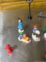 Silver table display of seven Christmas village figurines, a red fire hydrant, black street lamp, and a small toy vehicle.