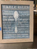 Rectangular wooden kitchen sign with table rules text in white on distressed blue background, framed in light wood