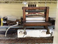 Front view of the antique wringer mounted on a white base, showing the wooden frame, hand crank, and metal clamps.