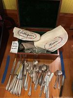 Wooden case open with various silver-plated flatware arranged, fabric pouches labeled for knives and tablespoons visible.