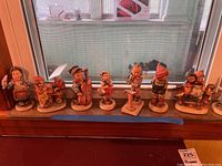 Photo showing a lineup of 9 Hummel figurines on a windowsill illustrating a variety of children in different poses including musical instruments and dancing.