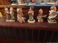 Six vintage Hummel figurines on display, various children posed with books, instruments, and objects.