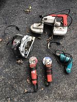 Group of six portable power tools including circular saw, band saw, angle grinders, and drill laid out on rough pavement.
