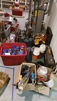 Overview of lot items including plastic tubs, jars, power tools, and lights arranged on basement floor.