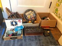 Wide angle photo showing many vintage radio-related items including boxes of tubes and crystals, a large vintage electrical component with control knobs, and additional boxes of parts on a carpeted floor.