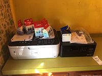 Two printers side by side: white Canon PIXMA MX330 with ink cartridge boxes on top, and black Epson XP-600 Expression Premium printer.