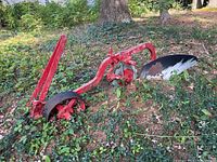 Wide view of the red metal plow attachment lying on ground partly overgrown by plant life showing full length and structure including blade and wheel.