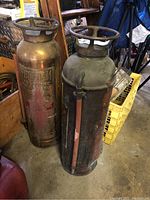 Two vintage cylindrical Firefoam fire extinguishers standing side by side, both showing surface rust and wear, stored indoors.