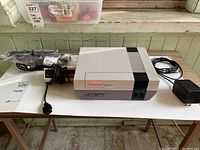 Front and cables view of the Nintendo NES console set laid out on white table, shows console, controller, power adapter, and AV cable.