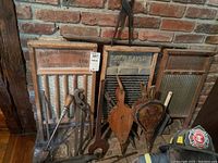 Photo showing 3 vintage washboards of varying sizes with wooden frames and metal scrubbing surfaces, wooden bellows, large wrench, pliers, and other hand tools grouped together against a brick wall.
