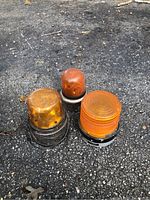 Three vintage amber warning lights placed on asphalt surface, showing different sizes and styles with visible wear and cables.