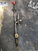 Full view of John Deere multi-tool with Toro trimmer attachment and weed whacker attachment (not shown) lying on asphalt surface with some other items in background
