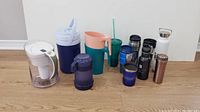 Photo showing 11 various drink containers of different materials and colors arranged on wooden floor against white wall